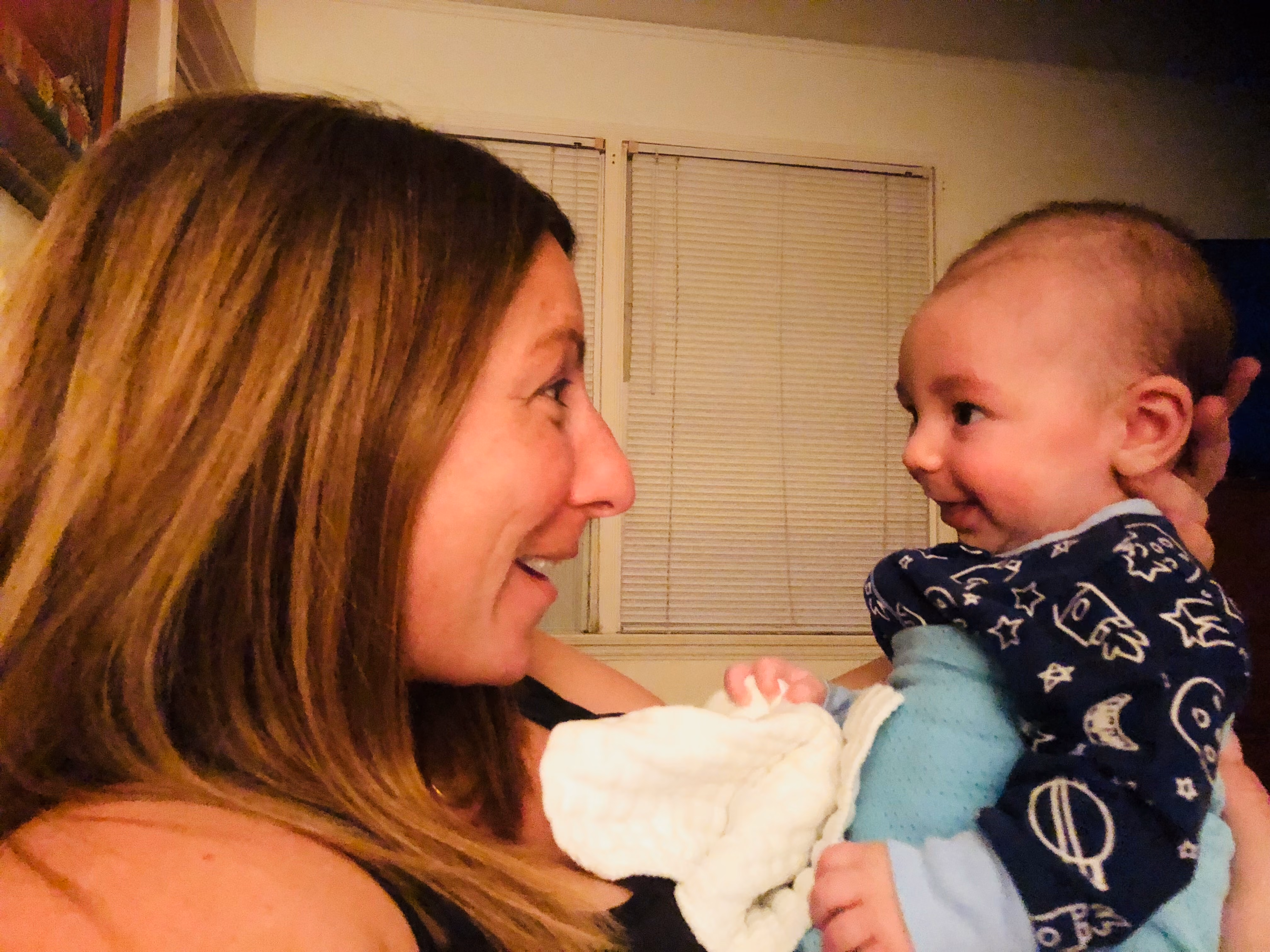A mother and her baby face to face, sharing a big smile in soft window light.